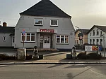 Restaurant Lotus Blick auf die Vorderseite des Restaurants. Es ist weiß-grau und liegt im Stadtgebiet. Zu sehen sind einige Häuser in der NÄhe..